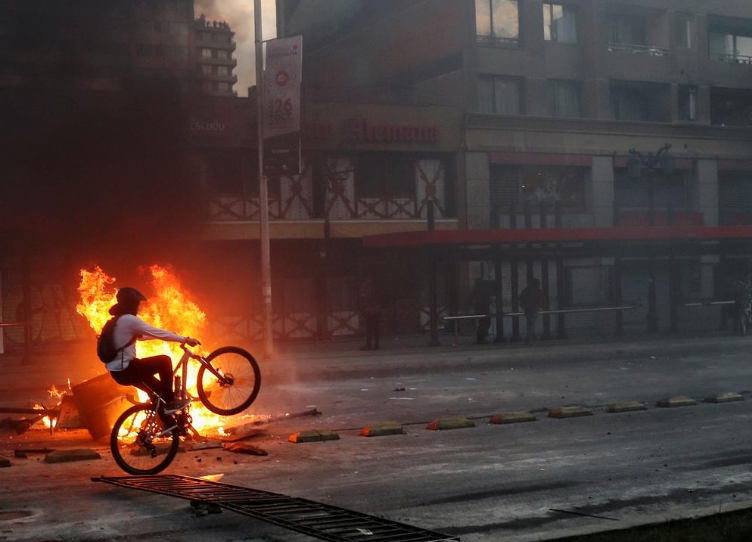 En la imagen de Reuters, un manifestante monta una bicicleta frente a una hoguera improvisada durante una protesta en Santiago de Chile.