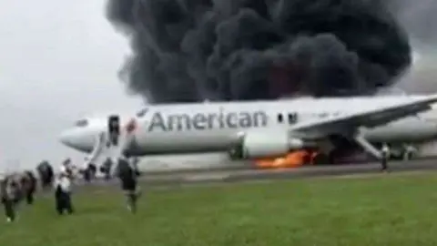 Pasajeros corren por la pista tras abandonar el avión en llamas de American Airlines. (Twittter)