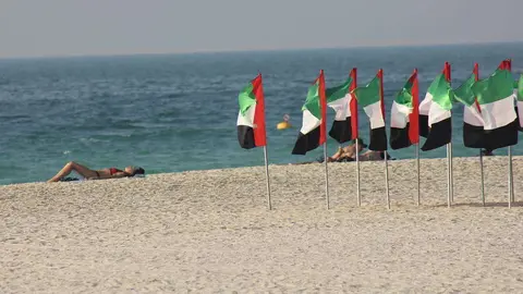 Bañistas toman el sol en la playa de Kite Beach en Dubai junto a banderas de Emiratos Árabes que ondean al viendo. (R. Pérez / EL CORREO)