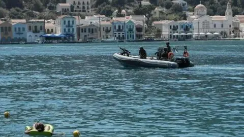 Una mujer toma el sol en el mar, mientras que una embarcación militar griega se dirige hacia la isla de Kastelórizo, epicentro de las disputas entre Grecia y Turquía. (AFP)