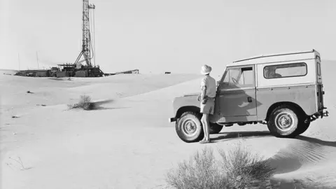 Un trabajador con su Land Rover observa una plataforma de perforación en el campo petrolero de Murban Bab en 1964. (The Naional)